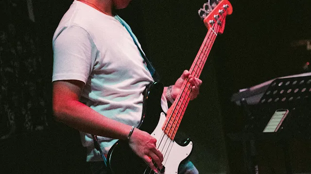 O Ritmo do Baixo no Palco Fotografia em plano médio e de perfil de uma pessoa a tocar um baixo elétrico num palco escuro. A pessoa veste uma t-shirt branca simples e o baixo tem um corpo preto com um escudo branco e uma cabeça vermelha. A mão esquerda pressiona as cordas no braço do instrumento, enquanto a mão direita está posicionada sobre as cordas perto dos captadores. O ambiente tem uma iluminação contrastante com tons avermelhados, e ao fundo, do lado direito, é possível ver parte de um suporte com uma pauta ou tablet.