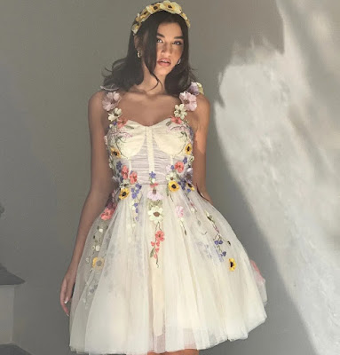 Whimsical Garden Guest Look: 3D Floral Appliqué Mini Dress A young woman posing in a cream-colored, tulle mini dress featuring vibrant 3D floral appliqués and embroidery. She is wearing a matching floral headband that complements the whimsical and romantic aesthetic, perfect for a creative or informal garden wedding.