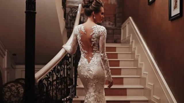 Vestido de noiva rendado com costas abertas elegantes Noiva a subir escadas com vestido de noiva rendado, mangas compridas e costas abertas, num ambiente clássico e elegante