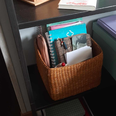 Cesta de Organização no Escritório Cesta de palha colocada numa prateleira de escritório, contendo cadernos, blocos, estojos e documentos organizados.