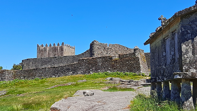 Lindoso: Entre o Castelo e os Espigueiros Vista do Castelo de Lindoso com as muralhas de pedra ao fundo e um espigueiro tradicional em primeiro plano num dia de céu azul.