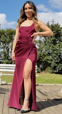 Look de Convidadas: Aposta no Vermelho Borgonha para um Visual Sophisticado Foto de corpo inteiro de uma mulher posando em um deck de madeira ao ar livre. Ela usa um vestido longo na cor vermelho borgonha, com decote reto, drapeado na cintura e uma fenda lateral profunda. Ela calça sapatos de salto alto pretos com detalhes brilhantes no tornozelo e na ponta.