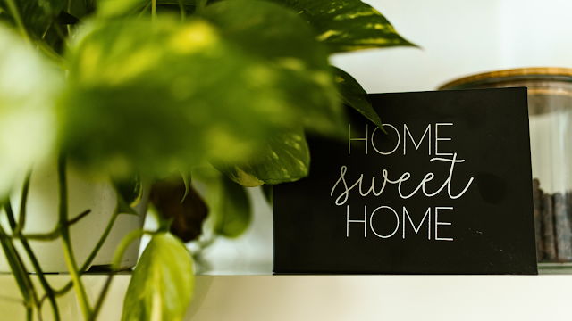 Home Sweet Home – Aproveitar o tempo em casa Placa decorativa com a frase "Home Sweet Home" ao lado de plantas verdes, simbolizando conforto e bem-estar em casa.