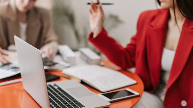 Reunião de trabalho com comunicação assertiva e colaboração Duas mulheres em reunião profissional, uma a comunicar de forma assertiva enquanto aponta com uma caneta, com computador portátil, caderno e telemóvel sobre a mesa