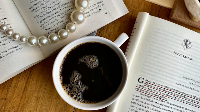 Momentos de Calma e Inspiração Clássica Vista superior de uma mesa de madeira clara com uma xícara de café preto no centro. Ao redor da xícara, dois livros abertos com textos impressos e um colar de pérolas clássico repousando sobre as páginas de um deles. A iluminação é suave e natural, criando uma composição elegante e intelectual que remete a um momento de estudo ou lazer tranquilo.