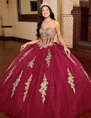 Realeza em Borgonha e Ouro: O Vestido dos Sonhos para Quinceañera Uma imagem cativante de uma jovem deslumbrante vestindo um opulento vestido de Quinceañera. O vestido, em um rico tom borgonha, apresenta um corpete sem alças coberto por intrincados bordados dourados. A saia de baile volumosa é feita de camadas de tule brilhante e é acentuada por apliques dourados verticais que descem em cascata. Ela usa uma coroa de cristal cintilante e joias de declaração, incluindo um colar e brincos reluzentes. Seu cabelo escuro e ondulado flui pelas costas enquanto ela sorri radiantemente, irradiando confiança e alegria em um ambiente elegante de salão de festas.