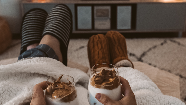 O refúgio perfeito: sofá, mantas e a minha maratona de eleição. Perspetiva em primeira pessoa de duas pessoas sentadas num sofá, cobertas por uma manta felpuda branca. Ambas seguram canecas de vidro com uma bebida cremosa (café tipo dalgona) nas mãos. Vêem-se as pernas esticadas, uma com meias às riscas e calças de pijama xadrez, e a outra com meias de lã castanhas. Ao fundo, uma sala de estar com uma televisão desligada sobre um móvel branco, criando um ambiente de conforto e relaxamento.