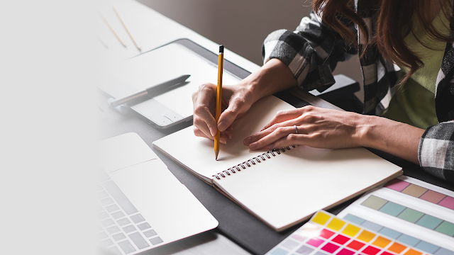 Pessoa escrevendo em caderno de anotações, com um laptop e paletas de cores ao lado, em uma mesa de trabalho criativa.