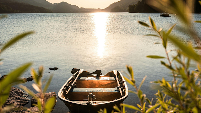 Amanhecer Dourado no Lago ou Barco a Remo ao Pôr do Sol Fotografia em close-up de um pequeno barco a remo de metal estacionado à beira de um lago tranquilo durante o pôr do sol. A luz dourada do sol reflete intensamente na água, criando um rastro brilhante. A cena é emoldurada por folhas verdes desfocadas em primeiro plano, com montanhas suaves ao fundo sob um céu claro de fim de tarde.