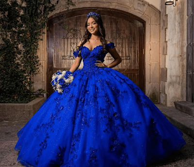 Realeza em Azul Royal: O Deslumbrante Vestido de Quinceañera Uma imagem deslumbrante de uma jovem posando para sua Quinceañera. Ela veste um opulento vestido de baile azul royal com um corpete sem alças e uma saia volumosa, cobertos por intrincados apliques florais e glitter cintilante. Ela usa uma tiara de cristal reluzente e brincos combinando, e segura um buquê de mixed rosas brancas e azuis. O cenário é um pátio de pedra rústico com grandes portas de madeira arqueadas, iluminado por lanternas de parede quentes e adornado com trepadeiras verdes.