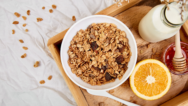 Taça de cereais integrais com passas, acompanhada de leite, meia laranja, mel e compota, num tabuleiro de madeira sobre um lençol branco – sugestão de pequeno-almoço saudável.