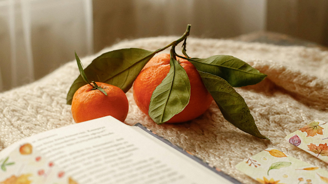 Leitura aconchegante com toque de outono Fotografia de tangerinas frescas com folhas verdes sobre uma manta bege, ao lado de um livro aberto e marcadores com desenhos de folhas de outono, criando um ambiente acolhedor e outonal.