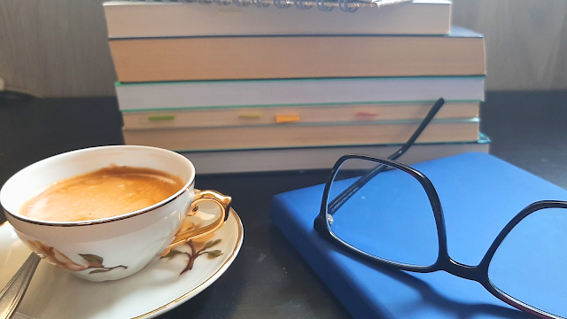 magem de uma secretária com livros empilhados, uma chávena de café e óculos, criando um ambiente de trabalho aconchegante e produtivo.