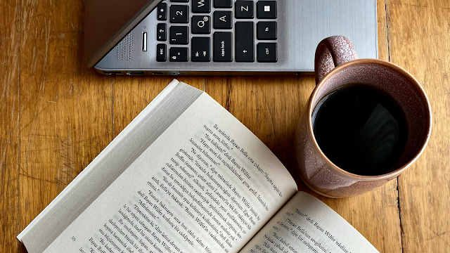 poder das pequenas pausas: leitura, café e inspiração Livro aberto ao lado de um portátil e de uma chávena de café sobre uma mesa de madeira, simbolizando um momento de leitura e tranquilidade.