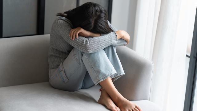 Mulher em sofrimento, símbolo da violência contra as mulheres Mulher sentada num sofá, abraçando os joelhos e escondendo o rosto, representando o impacto emocional da violência contra as mulheres e da falta de consentimento