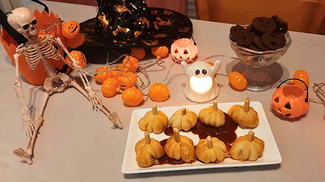 Jantar de Halloween com mini pizas em forma de abóbora Mesa decorada para um jantar de Halloween, com mini pizas em formato de abóbora servidas num prato branco, decoração temática com esqueleto, abóboras luminosas, chapéu de bruxa, luzes decorativas e doces de chocolate.