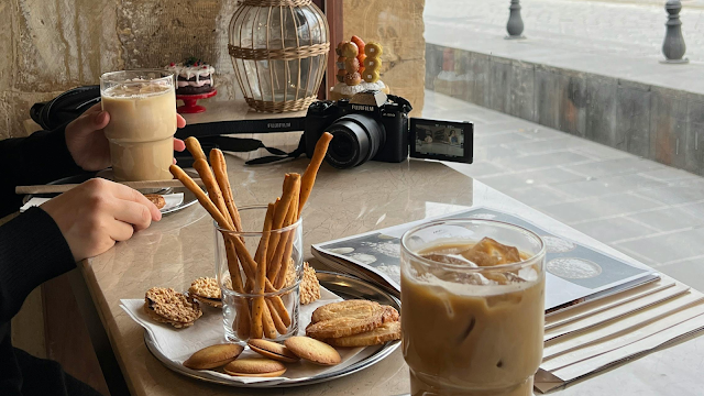 Cafés, Inspiração e Pequenos Prazeres de Janeiro Mesa de café com bolachas, grissinis e café gelado ao lado de uma câmara fotográfica, simbolizando momentos inspiradores e criativos de janeiro.
