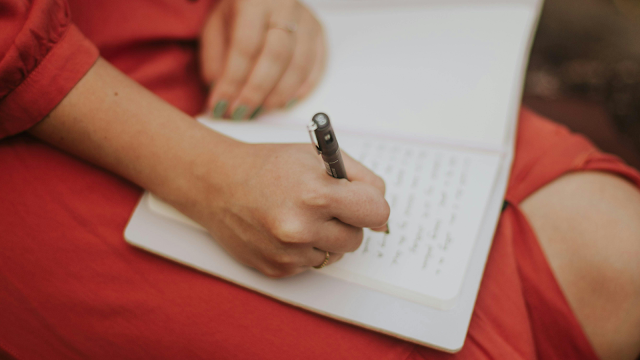 Mulher escrevendo em um caderno. Close-up das mãos de uma pessoa com vestido vermelho escrevendo com uma caneta preta em um caderno de páginas brancas apoiado no colo.