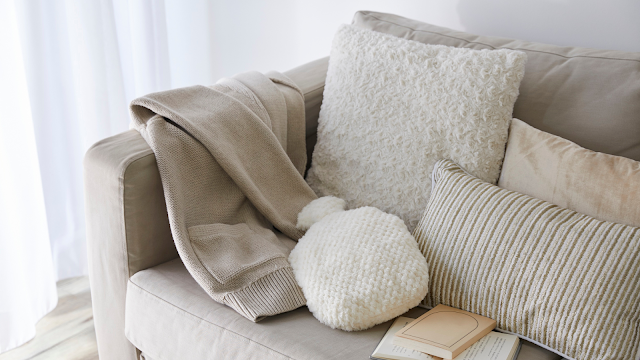 Conforto e tranquilidade em casa – Inspiração Soft Living Sofá bege com almofadas macias, manta de malha e livro aberto, criando um ambiente acolhedor de soft living.