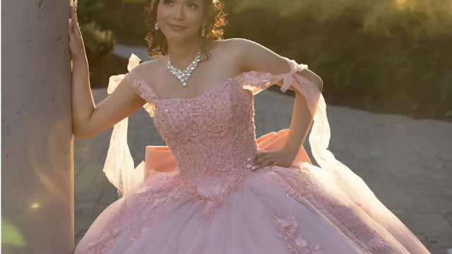 Uma Noite de Princesa: A Magia da Quinceañera Uma jovem radiante com um deslumbrante vestido de Quinceañera rosa pastel. O corpete é rendado e brilhante, e as alças são fluidas e delicadas. Ela usa joias reluzentes e seu cabelo está penteado com carinho. Ela se apoia em uma coluna, olhando para o horizonte com um sorriso sonhador, enquanto a luz dourada do sol poente a envolve, criando uma atmosfera mágica e inesquecível.