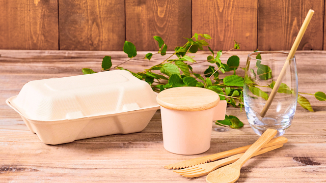 Produtos ecológicos reutilizáveis em madeira e bambu Conjunto de produtos ecológicos reutilizáveis, incluindo talheres de bambu, copo com palhinha e caixa de comida biodegradável sobre uma mesa de madeira.