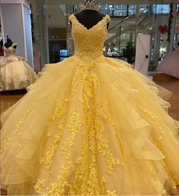 Sol e Sofisticação: O Vestido de Quinceañera Amarelo e Ouro Uma imagem deslumbrante de um vestido de Quinceañera em um tom radiante de amarelo sol, exibido em um manequim em um elegante salão de baile. O vestido apresenta um corpete sem alças com intrincados bordados dourados e apliques florais cintilantes. A volumosa saia de baile é feita de camadas de tule suave e é acentuada por babados estruturados e apliques dourados verticais que descem em cascata. O manequim usa uma coroa de cristal cintilante. O ambiente inclui painéis de parede dourados, espelhos altos que refletem o ambiente e grandes árvores de cerejeira artificiais com flores rosa suave, criando um contraste dramático e sofisticado. A luz quente do lustre ilumina a cena.
