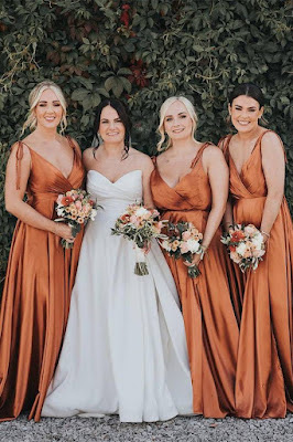 Inspiração de Cor: Vestidos Cobre para um Casamento Cheio de Charme Noiva vestida de branco posa sorridente ao lado de três damas de honor com vestidos longos de cetim na cor cobre, com alças de amarrar e decote em V, segurando bouquets de flores em tons quentes diante de um fundo de folhagem verde.