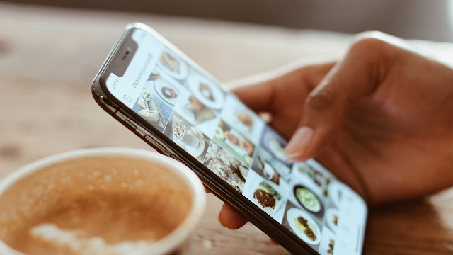 Navegar no Instagram com café Pessoa a segurar um smartphone enquanto navega no feed do Instagram, com fotografias de comida visíveis no ecrã, ao lado de uma chávena de café.