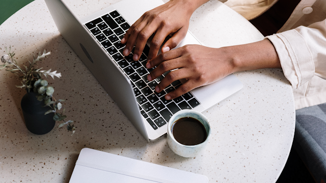 Escrever no blog com café e inspiração no dia a dia Mãos a escrever num computador portátil sobre uma mesa redonda, com uma chávena de café e uma pequena planta, num ambiente calmo e inspirador para criar conteúdos para um blog.