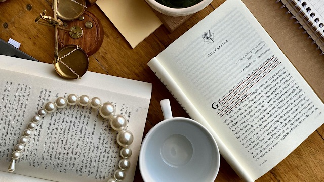 Filosofia no quotidiano: reflexões entre livros e café Livros abertos sobre uma mesa de madeira, acompanhados por uma chávena de café vazia, um colar de pérolas, um bloco de notas e óculos antigos, criando uma atmosfera de leitura e reflexão filosófica.