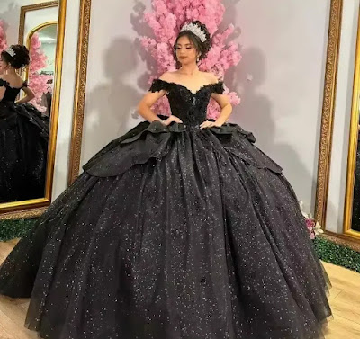 Uma imagem deslumbrante de uma jovem posando em um luxuoso salão de baile para sua Quinceañera. Ela veste um opulento vestido de baile preto com um corpete sem alças de renda intrincada e uma saia volumosa coberta por glitter cintilante que imita um céu estrelado. A saia apresenta sobreposições de babados estruturados. Ela usa uma tiara de cristal reluzente e brincos combinando. O cenário inclui painéis de parede dourados, espelhos altos que refletem o ambiente e grandes árvores de cerejeira artificiais com flores rosa suave, criando um contraste dramático e sofisticado. A luz quente do lustre ilumina a cena. Gótico Glamoroso: O Deslumbrante Vestido de Quinceañera Preto e Glitter