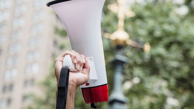 A Voz Que Todos Querem Ter Mão a segurar um megafone durante uma manifestação, simbolizando expressão pública e liberdade de opinião.