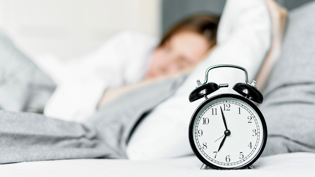 Mulher a dormir ao fundo, com um relógio em primeiro plano, simbolizando o início de uma nova rotina matinal.