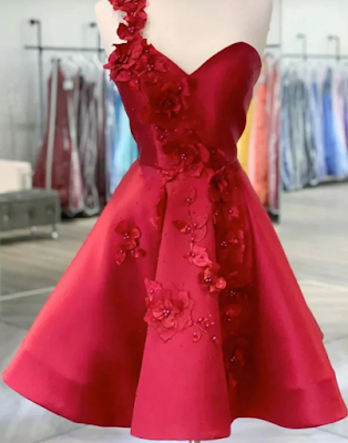 Red One-Shoulder Satin Dress with 3D Floral Appliqués A vibrant crimson red cocktail dress displayed on a white mannequin. The dress features a unique asymmetrical design with a single shoulder strap and a sweetheart neckline. It is heavily embellished with intricate 3D floral appliqués and matching red beads that cascade from the shoulder, down the bodice, and onto the voluminous A-line skirt. The fabric has a rich, satin-like sheen, and the background shows a blurred interior of a clothing boutique with various dresses on racks.