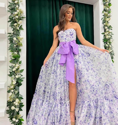 Lavender Floral Magic: Strapless Garden Wedding Guest Gown with Bow Accent A young woman posing in an elegant, strapless ball gown featuring a white fabric printed with delicate lavender-purple flowers. A large, solid lavender bow sash is cinched at the waist, and a dramatic high slit reveals her leg and nude heel. She stands against a backdrop of deep green curtains framed by beautiful floral garlands of white roses and greenery, showcasing a stunning, romantic look for a garden ceremony.