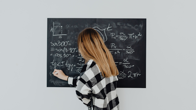 Matemática no quotidiano: fórmulas que explicam o mundo Pessoa a escrever fórmulas matemáticas num quadro preto, representando o ensino e a importância da matemática no dia a dia