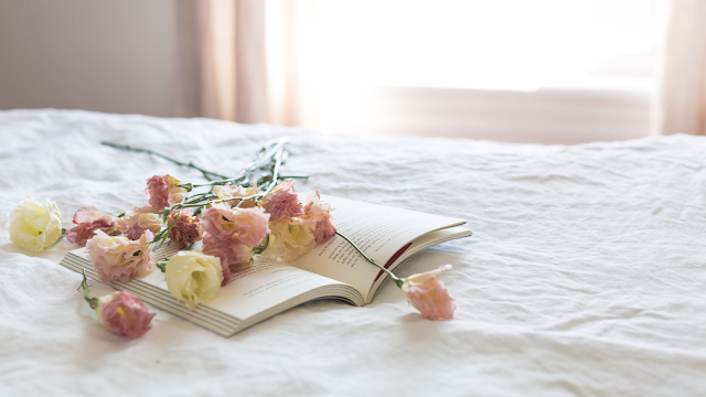 Flores delicadas sobre um livro aberto numa cama branca iluminada pela luz natural — momento de pausa e contemplação durante um apagão.