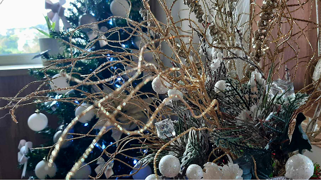 Brilho de Natal: Detalhes em Dourado e Branco Grande plano focado num arranjo decorativo de Natal composto por ramos finos e ondulados cobertos de purpurina dourada. No centro, destacam-se pequenas esferas brancas com textura de neve e cubos de acrílico transparentes. Ao fundo, de forma desfocada, vislumbra-se uma árvore de Natal verde decorada com luzes azuis e bolas brancas, criando um ambiente festivo e sofisticado.