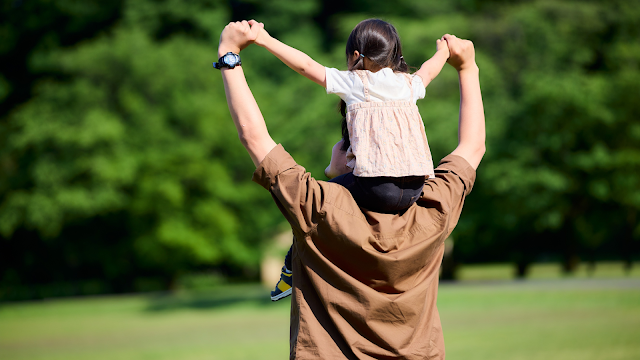Pai e filha a partilhar um momento ao ar livre Pai de costas com a filha aos ombros, de mãos dadas no ar, num parque verde num dia de sol