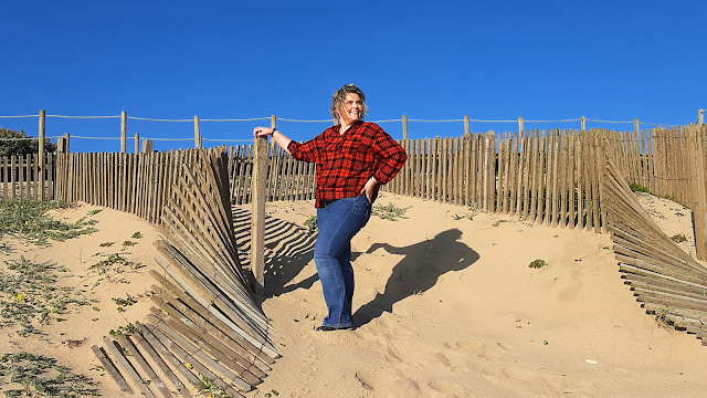 Mulher sorridente de camisa vermelha e calças de ganga posa nas dunas de areia sob um céu azul, junto a uma vedação de madeira — imagem descontraída em cenário de praia natural.