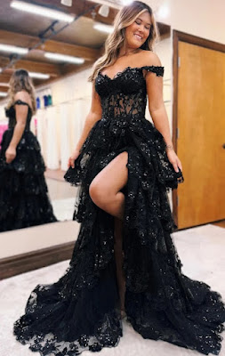 Jovem vestindo um deslumbrante vestido de festa preto com fenda e decote ombro a ombro Uma mulher jovem e sorridente com cabelo castanho ondulado está de pé num salão de noivas, posando para a câmara enquanto veste um luxuoso vestido de baile preto. O vestido tem um decote ombro a ombro adornado com renda e lantejoulas, um corpete de estilo corset em renda preta e uma saia de tule e renda em camadas com uma fenda alta que revela a sua perna esquerda e uma tatuagem no tornozelo. Ela está de pé sobre um tapete branco com o seu pé esquerdo apoiado numa cadeira oculta para mostrar a fenda do vestido. Ao fundo, vê-se um espelho grande que reflete a parte de trás do vestido e a loja com mais vestidos de festa e de noiva pendurados em cabides, e um teto alto com vigas expostas e luzes de pista. O chão é de azulejos claros.
