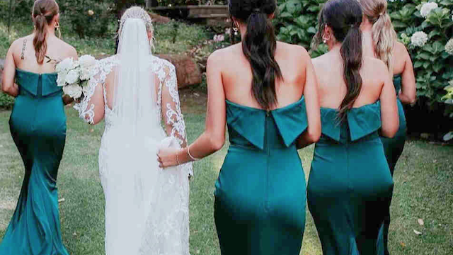 Damas de Honor em Verde Esmeralda: Elegância e Harmonia no Grande Dia Noiva caminhando num jardim com quatro damas de honor vestidas com vestidos longos de cetim na cor verde esmeralda, com decote sem alças e detalhe de laço nas costas, criando um visual elegante e coordenado.