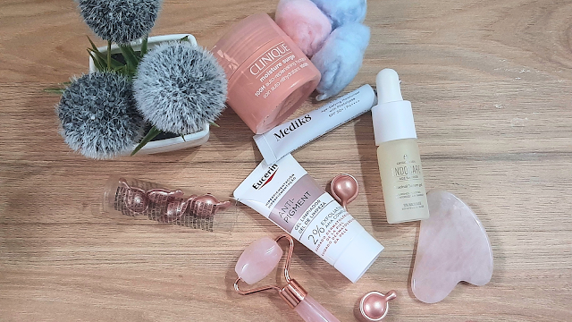 A Minha Rotina de Skincare com os Miminhos da Sweetcare Produtos de skincare sobre uma mesa de madeira, incluindo o Clinique Moisture Surge, o gel de limpeza Eucerin Anti-Pigment, o sérum Endocare Age Barrier, o protetor solar Medik8 e ferramentas de massagem facial em quartzo rosa.