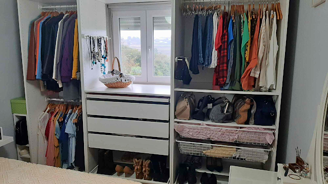 Arrumações de Outono: Closet Organizado e Funcional Fotografia de um closet organizado com roupa pendurada, gavetas brancas, sapatos arrumados em prateleiras e luz natural a entrar pela janela ao centro.