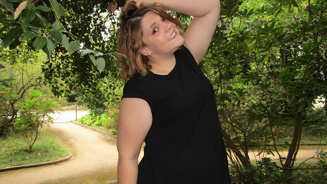 A Beleza de Aproveitar o Vosso Próprio Tempo Mulher a sorrir num jardim, com um vestido preto, posando de forma descontraída entre árvores e caminhos rodeados de natureza.