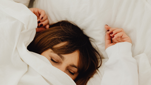 Mulher a dormir profundamente numa cama branca Mulher deitada na cama, tapada com um edredão branco, a dormir tranquilamente com expressão relaxada.