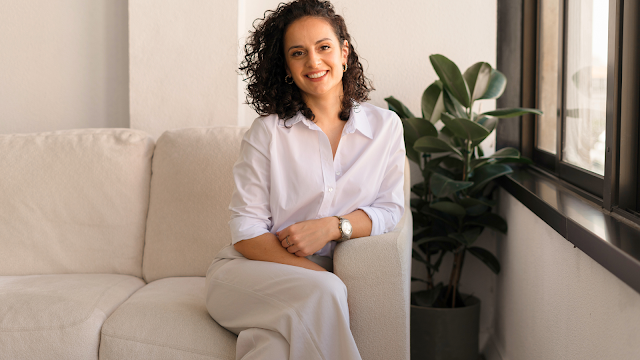 Joana Pereira: Marketing Digital com Propósito Mulher sentada num sofá claro, com camisa branca e calças claras, a sorrir para a câmara, junto a uma janela e uma planta decorativa.