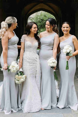 Elegância Simétrica: Vestidos Longos e Iguais para um Casamento Chic Noiva ao centro, vestida com um vestido de noiva rendado e estruturado, rodeada por três damas de honor que vestem vestidos longos iguais, de um ombro só, na cor cinza claro. Todas seguram bouquets de rosas brancas e sorriem num ambiente arquitetónico clássico.