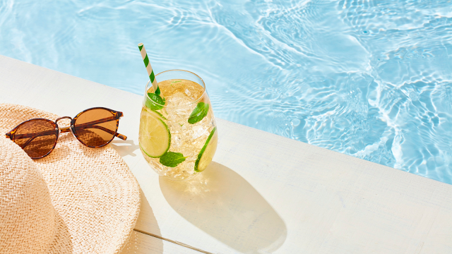 Dicas para aproveitar as férias de verão. Foto em close-up de uma mesa branca ao lado de uma piscina de águas azuis cristalinas. Sobre a mesa, há um copo com uma bebida gelada contendo rodelas de limão, folhas de hortelã e um canudo listrado em verde e branco. Ao lado, um par de óculos de sol de tartaruga e parte de um chapéu de palha, sugerindo um momento de descanso e lazer sob o sol.