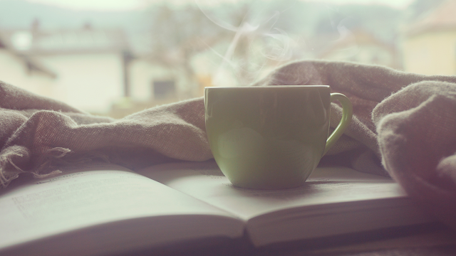Caneca de chá ou café fumegante sobre um livro aberto, com um tecido aconchegante enrolado ao fundo, transmitindo uma sensação de calma e conforto em um ambiente tranquilo.